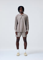 A person with dark, shoulder-length hair stands facing forward against a plain light background, wearing the STRIPE JERSEY set from HOMME PLISSÉ ISSEY MIYAKE with matching shorts, white socks, and beige shoes.