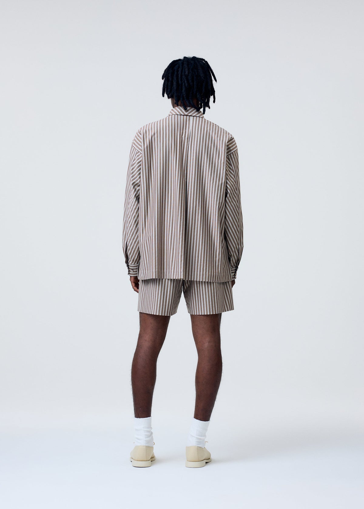 A person with dark, shoulder-length hair faces away, wearing the HOMME PLISSÉ ISSEY MIYAKE STRIPE JERSEY set—beige and white striped shirt with Italian collar and matching shorts—paired with white socks and beige shoes against a light background. view 7 of 8