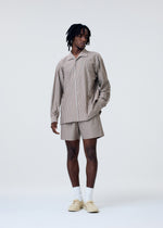 A person stands against a plain light background wearing the HOMME PLISSÉ ISSEY MIYAKE STRIPE JERSEY shirt and matching shorts, paired with white socks and cream-colored shoes, one hand in their pocket, looking to the side.