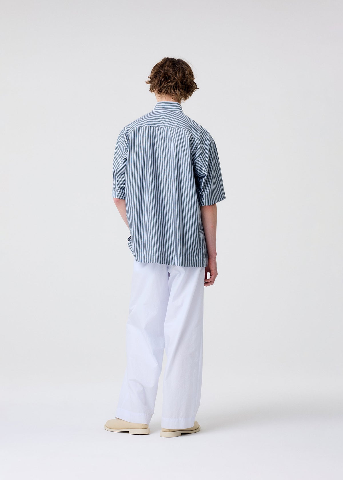 A person stands facing away from the camera, wearing the HOMME PLISSÉ ISSEY MIYAKE STRIPE JERSEY short-sleeved shirt with a standing collar, paired with loose white pants and beige shoes against a plain light background. view 7 of 9