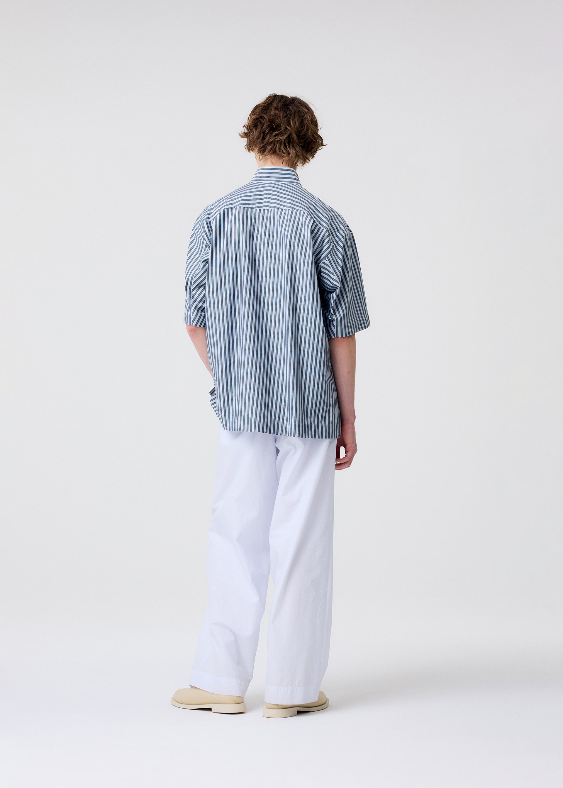 Zoomed view A person stands facing away from the camera, wearing the HOMME PLISSÉ ISSEY MIYAKE STRIPE JERSEY short-sleeved shirt with a standing collar, paired with loose white pants and beige shoes against a plain light background.