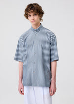 A young person with curly brown hair models the HOMME PLISSÉ ISSEY MIYAKE STRIPE JERSEY, a loose short-sleeved shirt with blue and white stripes and a standing collar, paired with white pants against a plain light background.