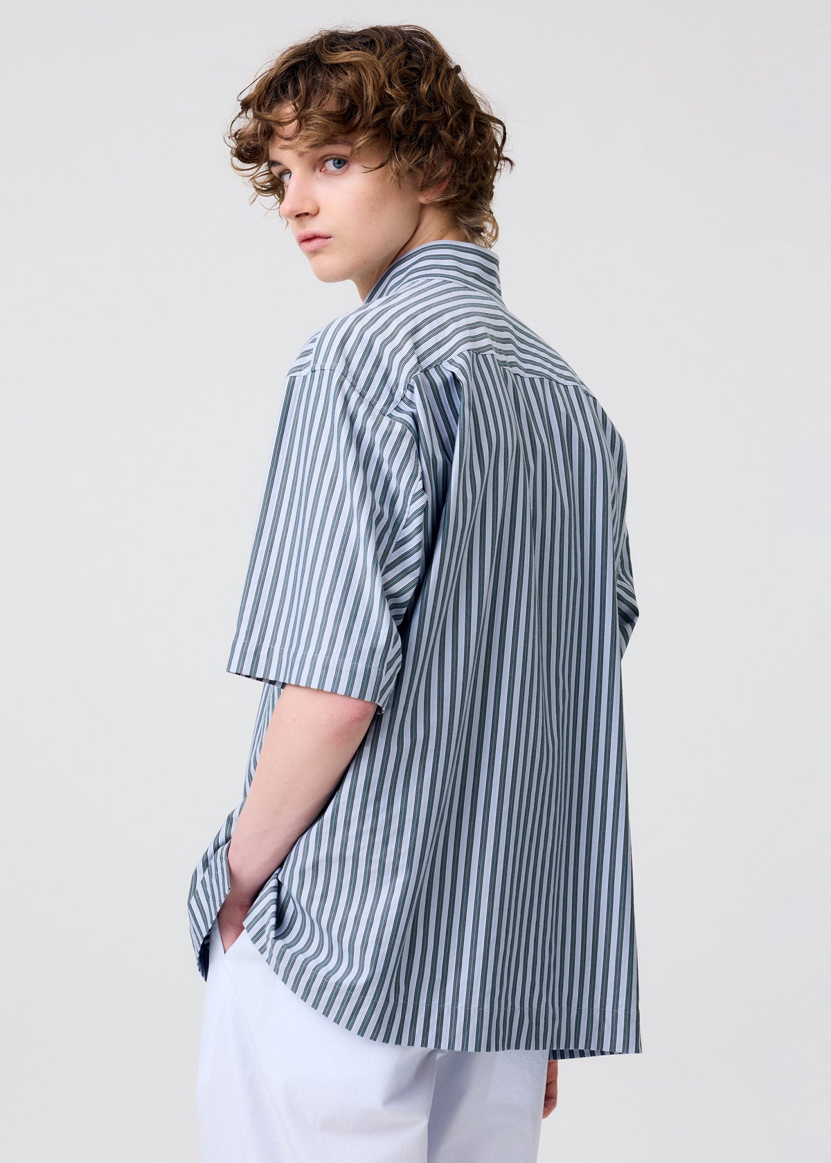 A person with curly brown hair models the HOMME PLISSÉ ISSEY MIYAKE STRIPE JERSEY—a loose, blue-and-white striped short-sleeved shirt—paired with white pants, standing slightly side-on and looking over their shoulder against a plain light background. view 9 of 9
