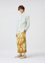 Wearing HOMME PLISSÉ ISSEY MIYAKE’s CRYSTALLINE JERSEY, a person with curly hair stands relaxed against a light background, layering the sheer long-sleeved shirt over loose, yellow-orange patterned pants for added depth.