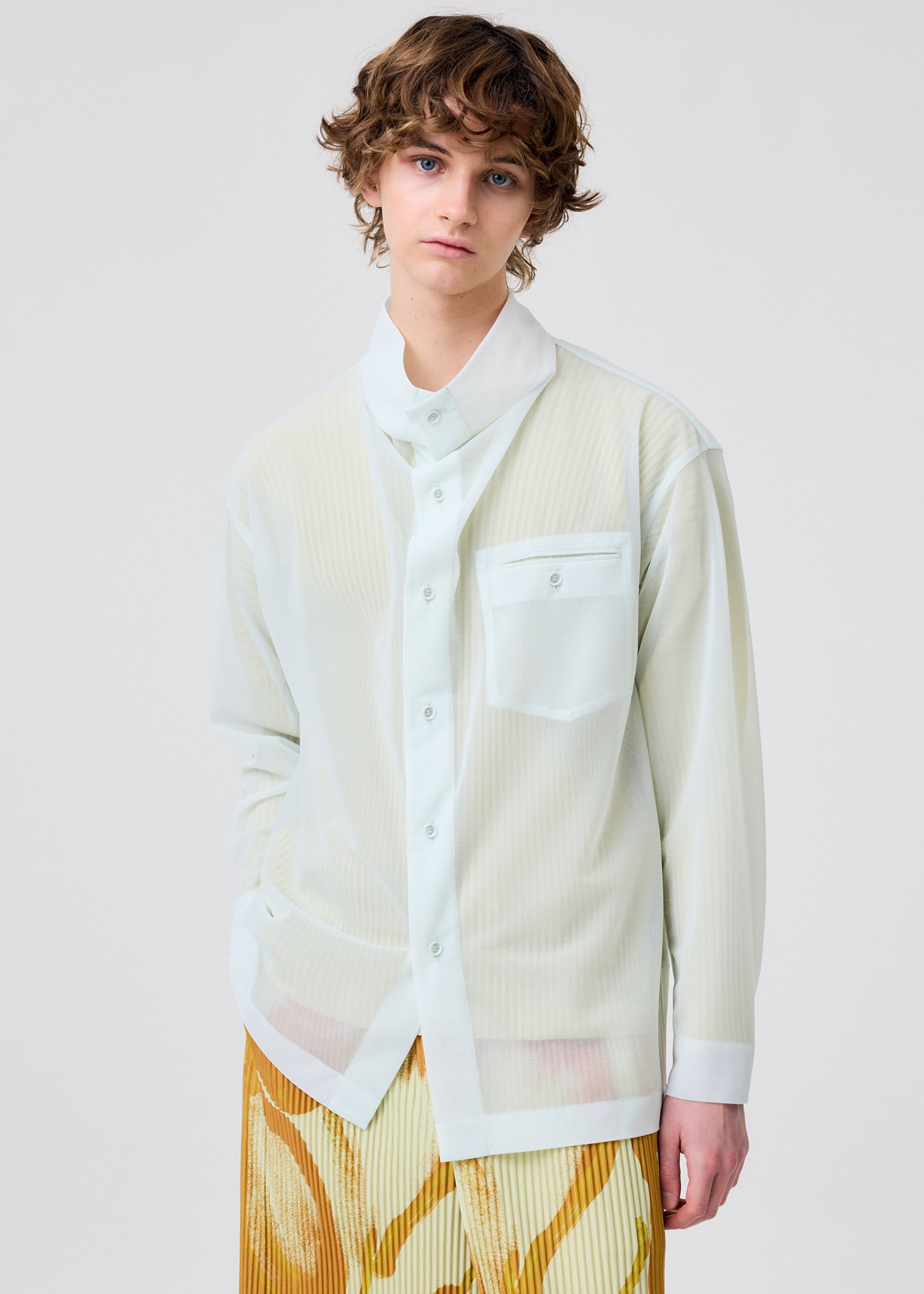 Zoomed view A person with wavy brown hair wears the CRYSTALLINE JERSEY by HOMME PLISSÉ ISSEY MIYAKE— a sheer, long-sleeved white button-up over a white collar and cuffs, paired with patterned yellow and white pants, against a plain light background.