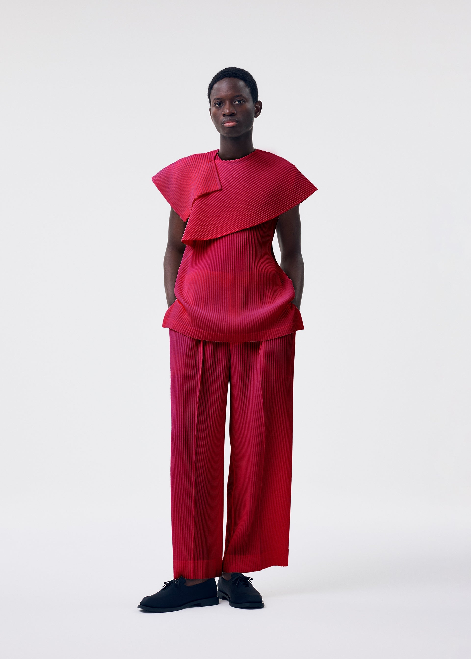 Zoomed view A person wears the MONTHLY COLORS : JANUARY set from HOMME PLISSÉ ISSEY MIYAKE—bold, structured red pleated wrap top with matching wide-legged pants and black shoes against a plain background.