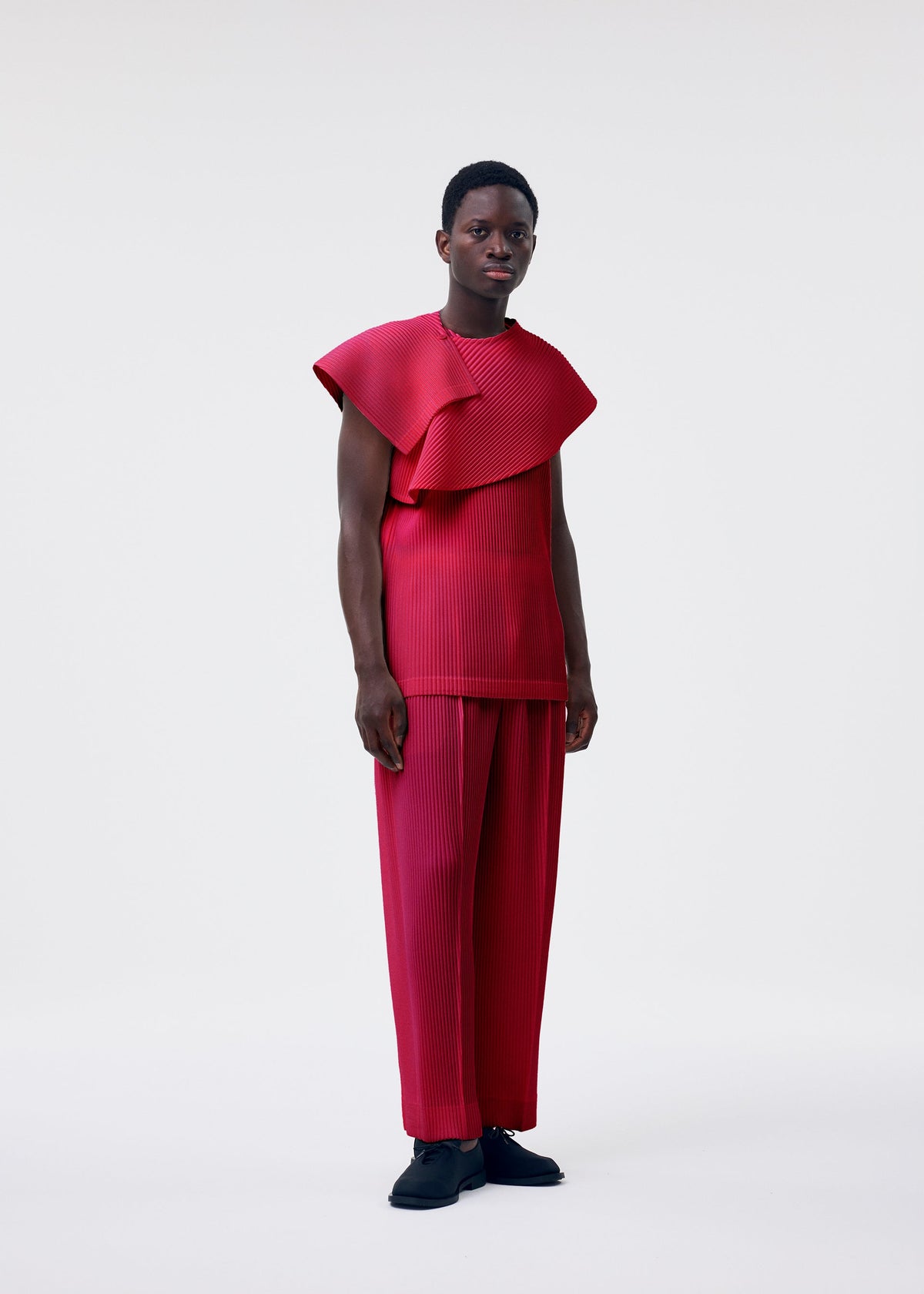 A person wears the HOMME PLISSÉ ISSEY MIYAKE MONTHLY COLORS : JANUARY set—a bright red scarf wrap top with sculptural shoulders and matching wide-leg pleated pants—against a plain white background, styled with short hair and black shoes. view 7 of 7