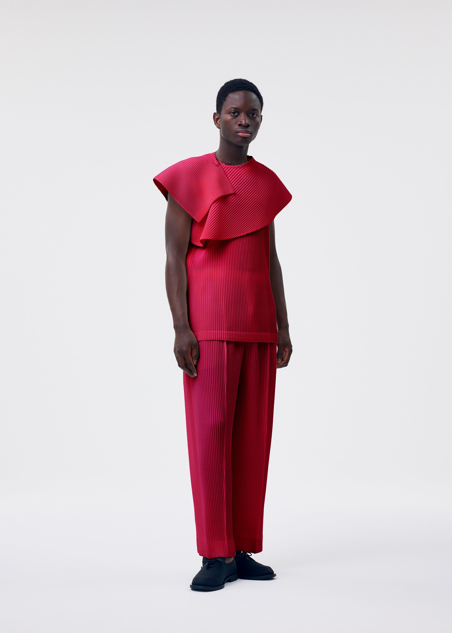 Zoomed view A person wears the HOMME PLISSÉ ISSEY MIYAKE MONTHLY COLORS : JANUARY set—a bright red scarf wrap top with sculptural shoulders and matching wide-leg pleated pants—against a plain white background, styled with short hair and black shoes.