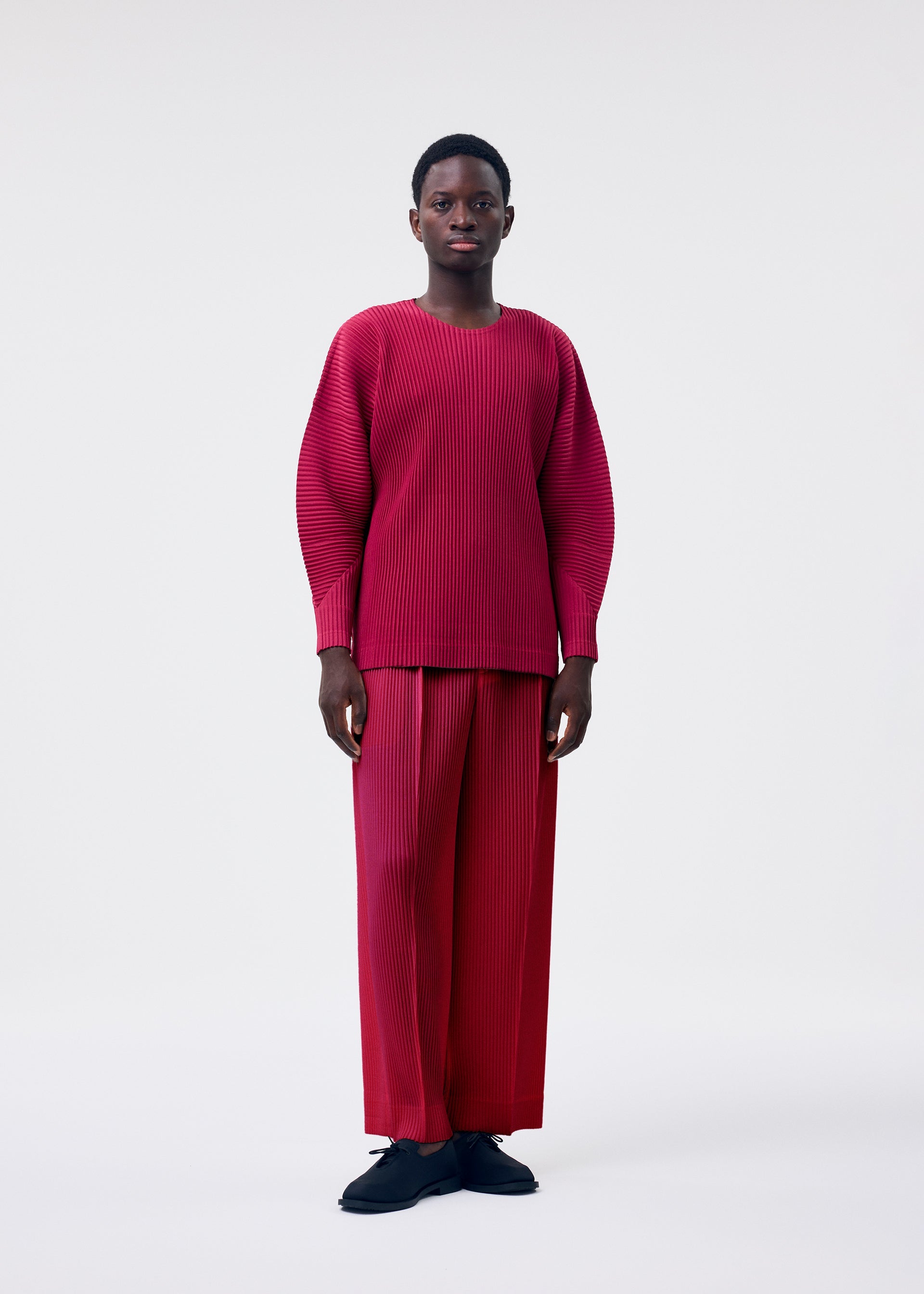 Zoomed view A person with short hair stands against a white background, wearing HOMME PLISSÉ ISSEY MIYAKE'S MONTHLY COLORS : JANUARY—a loose, lightweight red round-neck top and matching wide-leg ribbed pants—paired with black shoes.