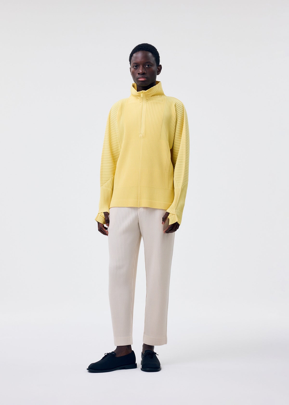 A person wearing the MONTHLY COLORS : MARCH yellow polyester long-sleeve top by HOMME PLISSÉ ISSEY MIYAKE, paired with cream pants and black shoes, stands against a white background, looking directly at the camera with a neutral expression. view 6 of 7
