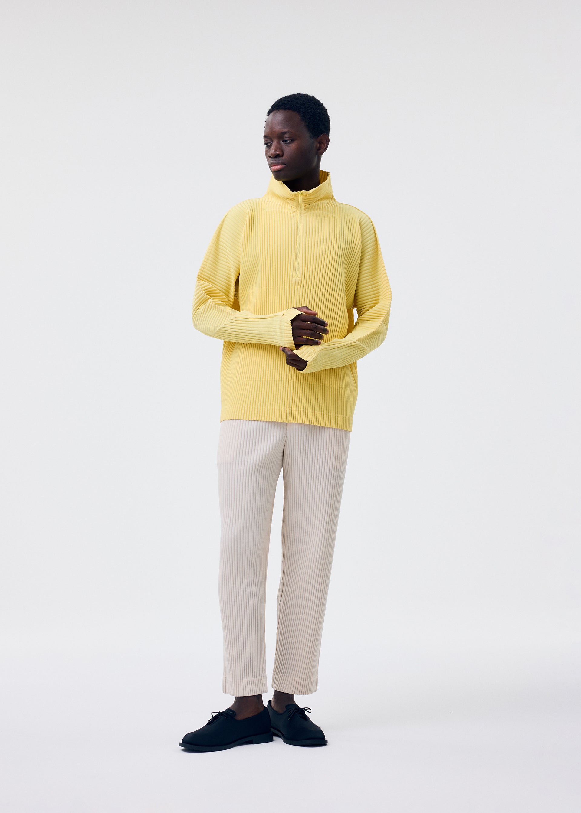 Zoomed view A person with short hair stands against a white background, wearing HOMME PLISSÉ ISSEY MIYAKE's MONTHLY COLORS : MARCH yellow ribbed polyester sweater with long sleeves and light beige pants, hands loosely clasped and looking to the side.