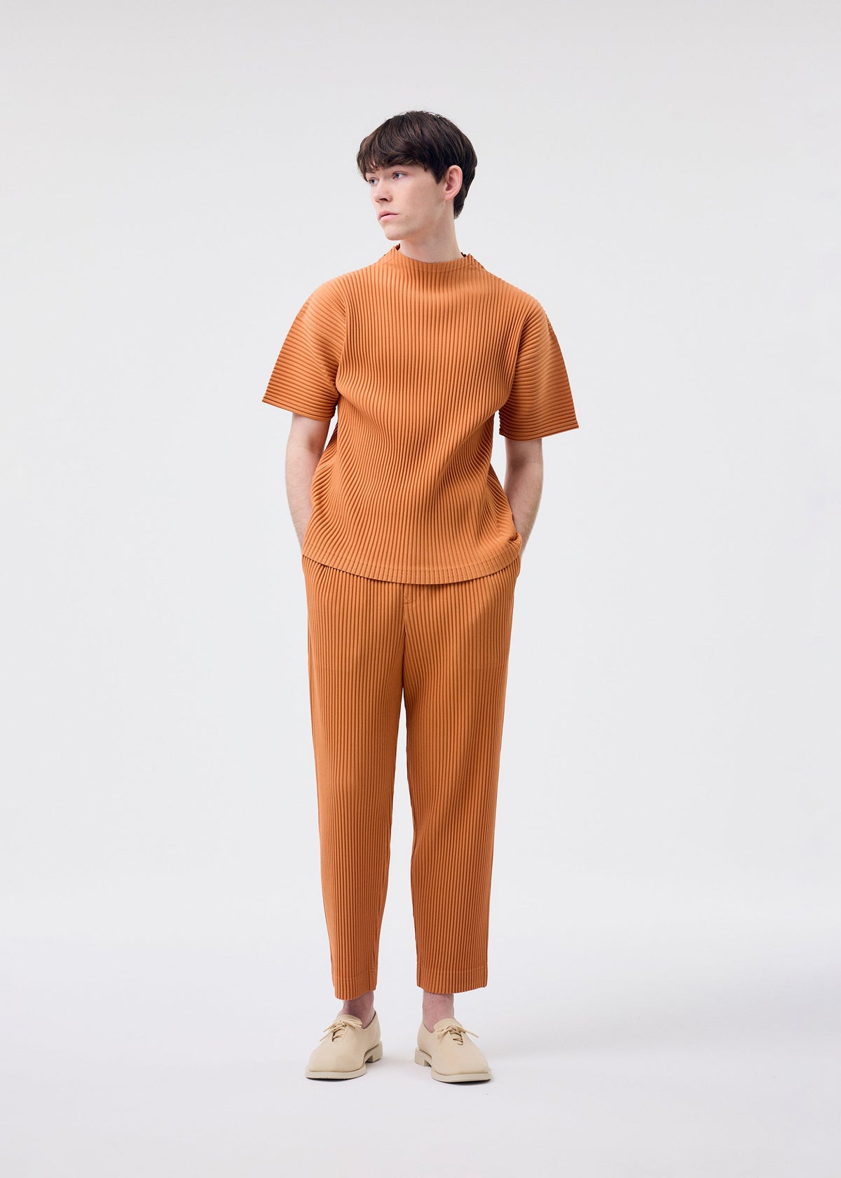 A young man, wearing the HOMME PLISSÉ ISSEY MIYAKE MONTHLY COLORS : MARCH light brown short-sleeved mock-neck top and pants set in quick-drying polyester, stands against a white background with his hands in his pockets and beige shoes, looking to the side. view 6 of 7