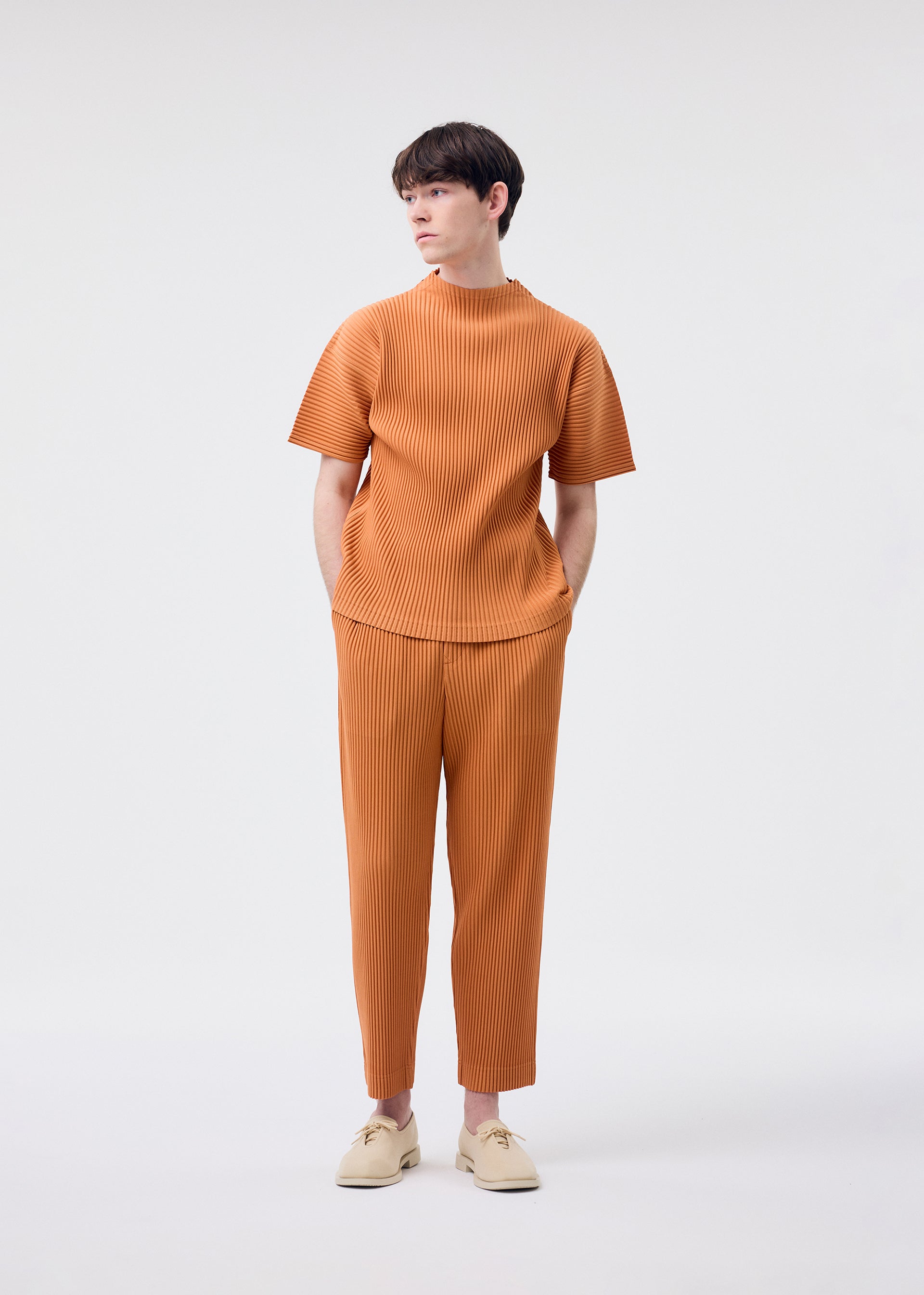 Zoomed view A young man, wearing the HOMME PLISSÉ ISSEY MIYAKE MONTHLY COLORS : MARCH light brown short-sleeved mock-neck top and pants set in quick-drying polyester, stands against a white background with his hands in his pockets and beige shoes, looking to the side.