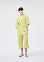 A person models the HOMME PLISSÉ ISSEY MIYAKE "MONTHLY COLORS : MAY" set—loose, light yellow round-neck top and matching wide-leg cropped pants—with pale green shoes, standing against a white background, hands in pockets.