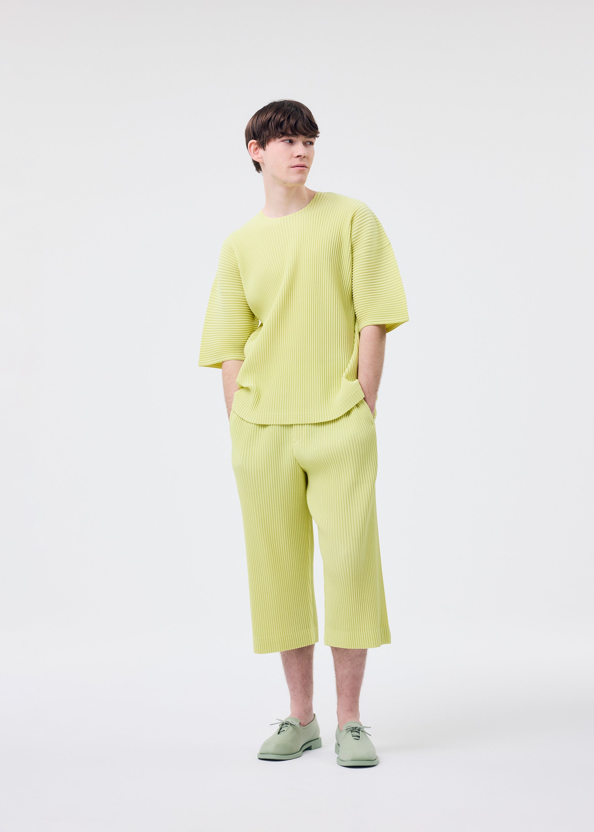 Zoomed view A person models the HOMME PLISSÉ ISSEY MIYAKE "MONTHLY COLORS : MAY" set—loose, light yellow round-neck top and matching wide-leg cropped pants—with pale green shoes, standing against a white background, hands in pockets.