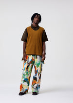 A person with short dreadlocks and a relaxed expression wears HOMME PLISSÉ ISSEY MIYAKE's MONTHLY COLORS : JUNE—featuring a brown short-sleeved shirt, mustard-yellow quick-drying sleeveless top, and wide-leg abstract-patterned pants.