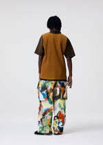 A person models the MONTHLY COLORS : JUNE collection by HOMME PLISSÉ ISSEY MIYAKE, featuring a brown ribbed polyester shirt and loose, abstract-print pants. The plain background highlights the vibrant, quick-drying outfits.