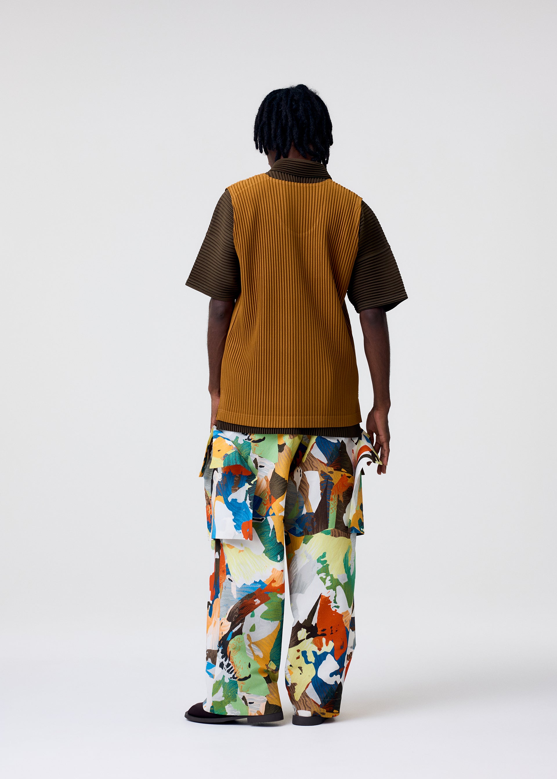 Zoomed view A person models the MONTHLY COLORS : JUNE collection by HOMME PLISSÉ ISSEY MIYAKE, featuring a brown ribbed polyester shirt and loose, abstract-print pants. The plain background highlights the vibrant, quick-drying outfits.