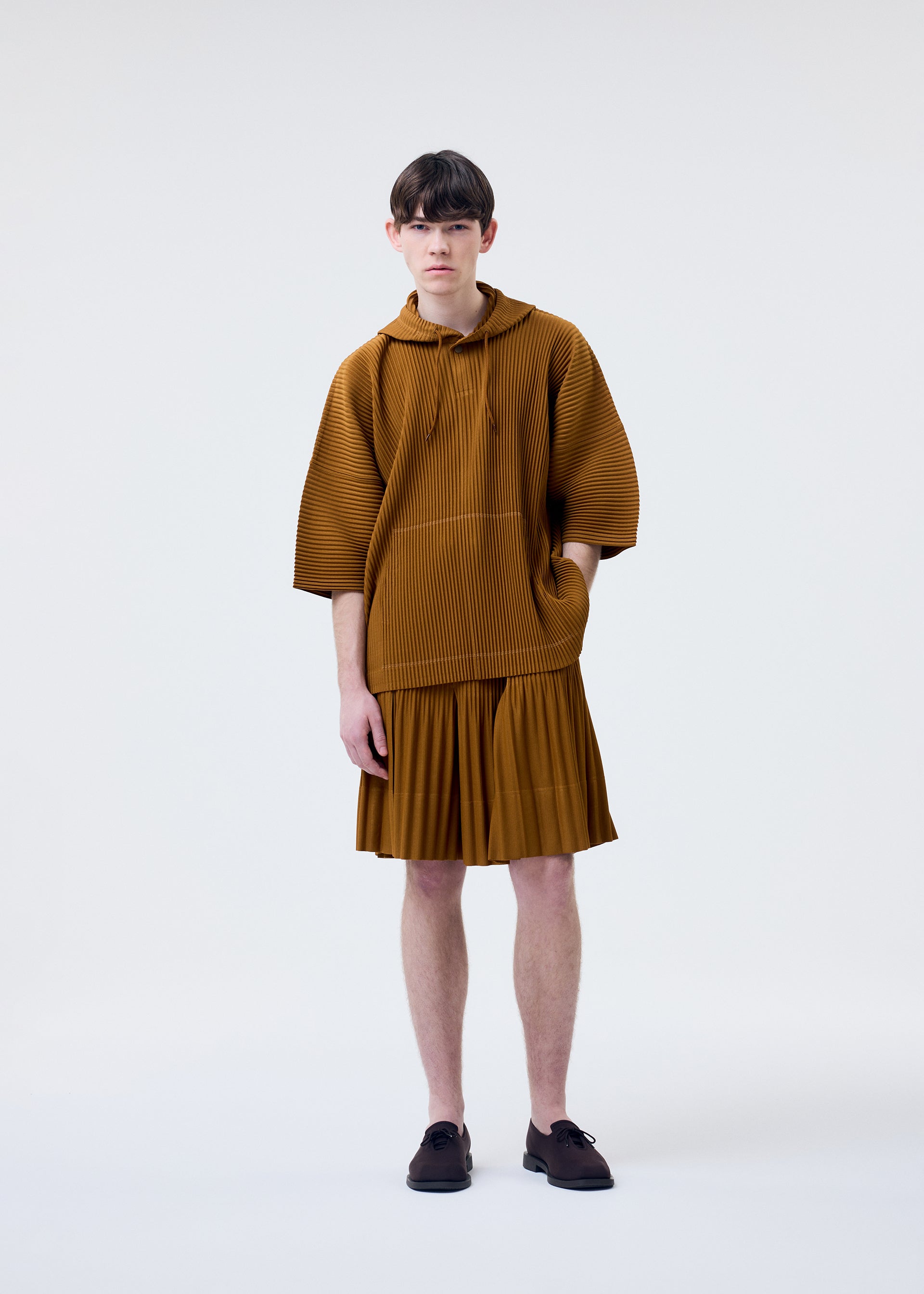 Zoomed view A person with short dark hair wears the HOMME PLISSÉ ISSEY MIYAKE MONTHLY COLORS : JUNE lightweight brown hooded pullover and matching pleated skirt, standing against a plain white background.