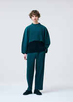A person with curly hair stands against a plain background, dressed in HOMME PLISSÉ ISSEY MIYAKE's COLOR PLEATS 1: a loose-fitting teal and black pleated jersey top with matching teal pleated pants, paired with black shoes.