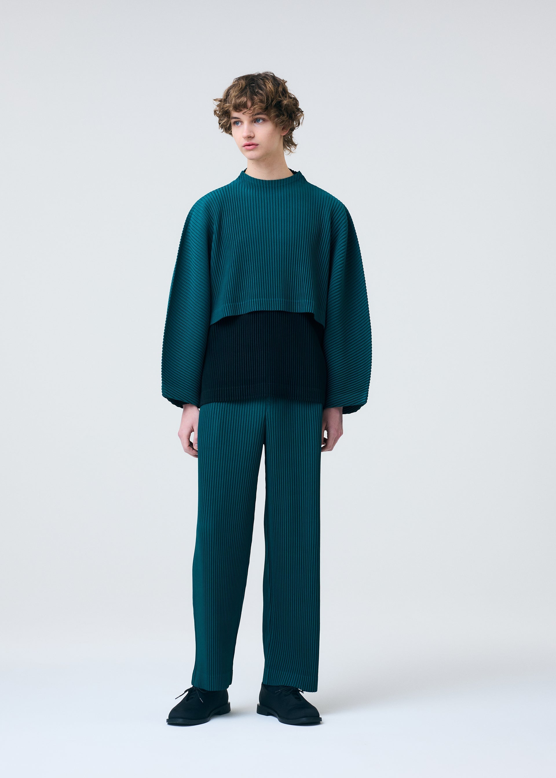 Zoomed view A person with curly hair wears HOMME PLISSÉ ISSEY MIYAKE’s COLOR PLEATS 1—a loose, teal and black pleated jersey top with matching teal pleated pants—standing in front of a plain light background, looking to the side with a relaxed posture.