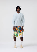 A person stands facing forward in a studio wearing the HOMME PLISSÉ ISSEY MIYAKE AGAVE MESH light blue knit long-sleeve top, colorful abstract shorts, cream socks, and beige shoes against a plain white background.