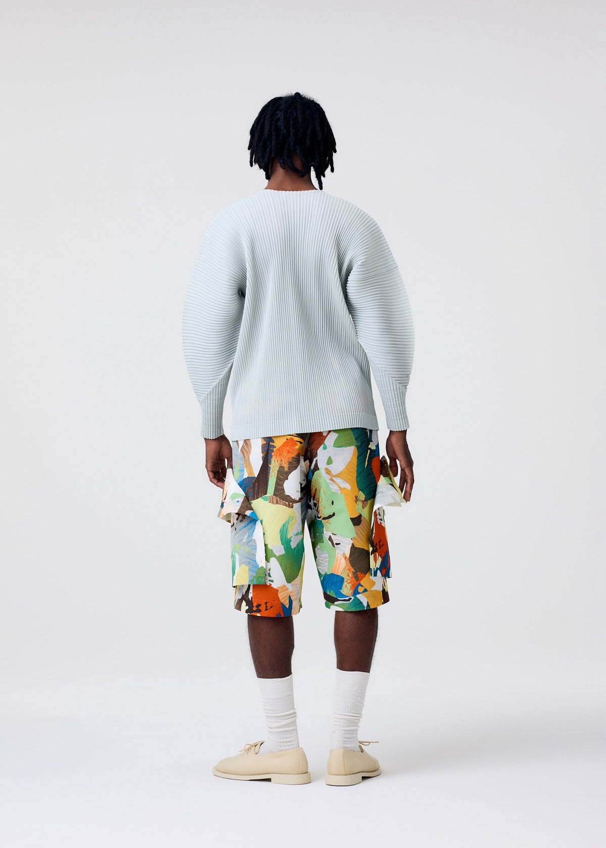 A person with short, dark dreadlocks stands facing away wearing the HOMME PLISSÉ ISSEY MIYAKE AGAVE MESH long-sleeve top, multicolored abstract shorts, white socks, and beige shoes against a plain white background. view 7 of 7