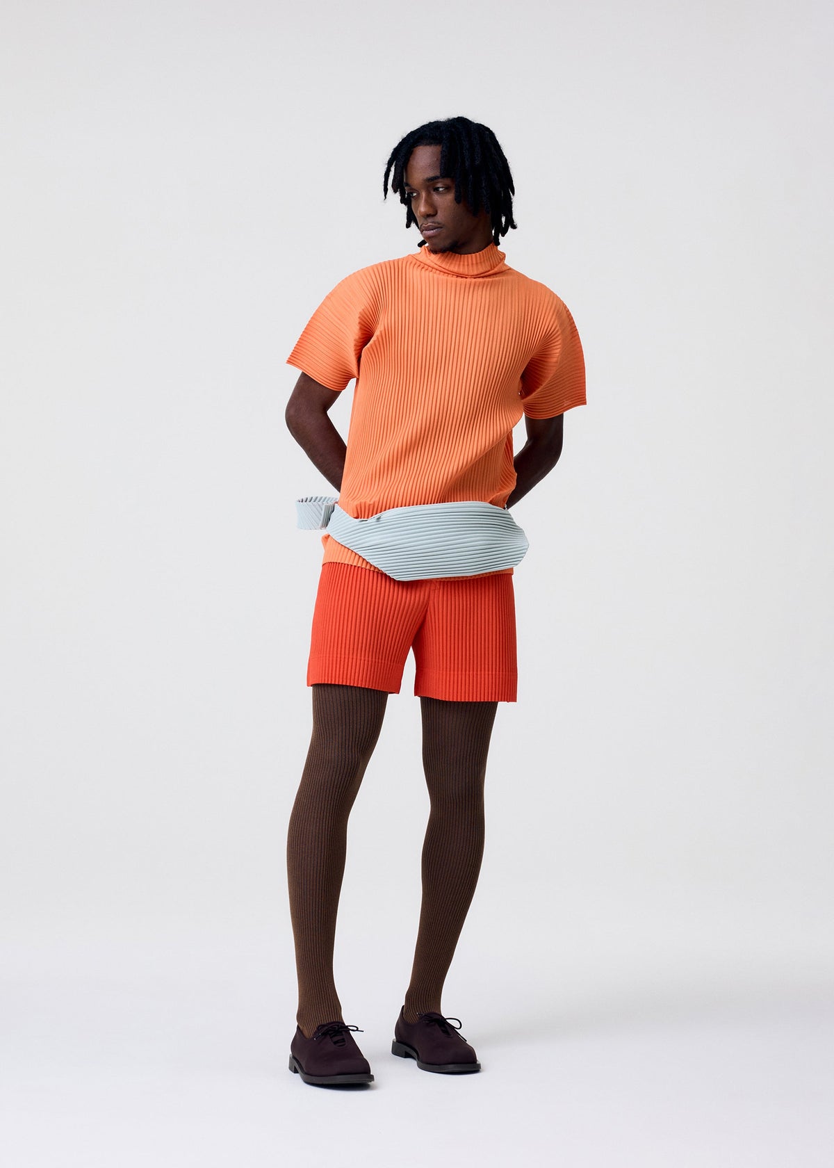 A person stands against a plain background, wearing the AGAVE MESH set—an orange ribbed high-neck top and matching shorts—by HOMME PLISSÉ ISSEY MIYAKE, with brown ribbed tights, dark brown shoes, and a pale blue textured belt bag. view 7 of 8