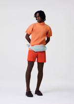 A person stands against a plain background, wearing the AGAVE MESH set—an orange ribbed high-neck top and matching shorts—by HOMME PLISSÉ ISSEY MIYAKE, with brown ribbed tights, dark brown shoes, and a pale blue textured belt bag.
