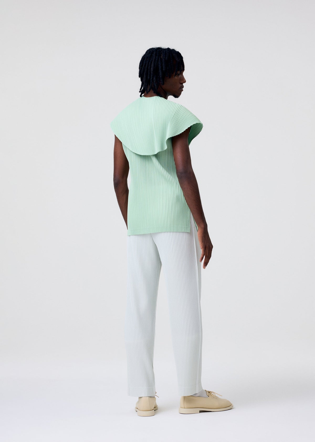 A person with short curly hair stands facing away, wearing the HOMME PLISSÉ ISSEY MIYAKE AGAVE MESH light green sleeveless knit top, light blue pleated pants, and beige shoes against a plain white background. view 6 of 7