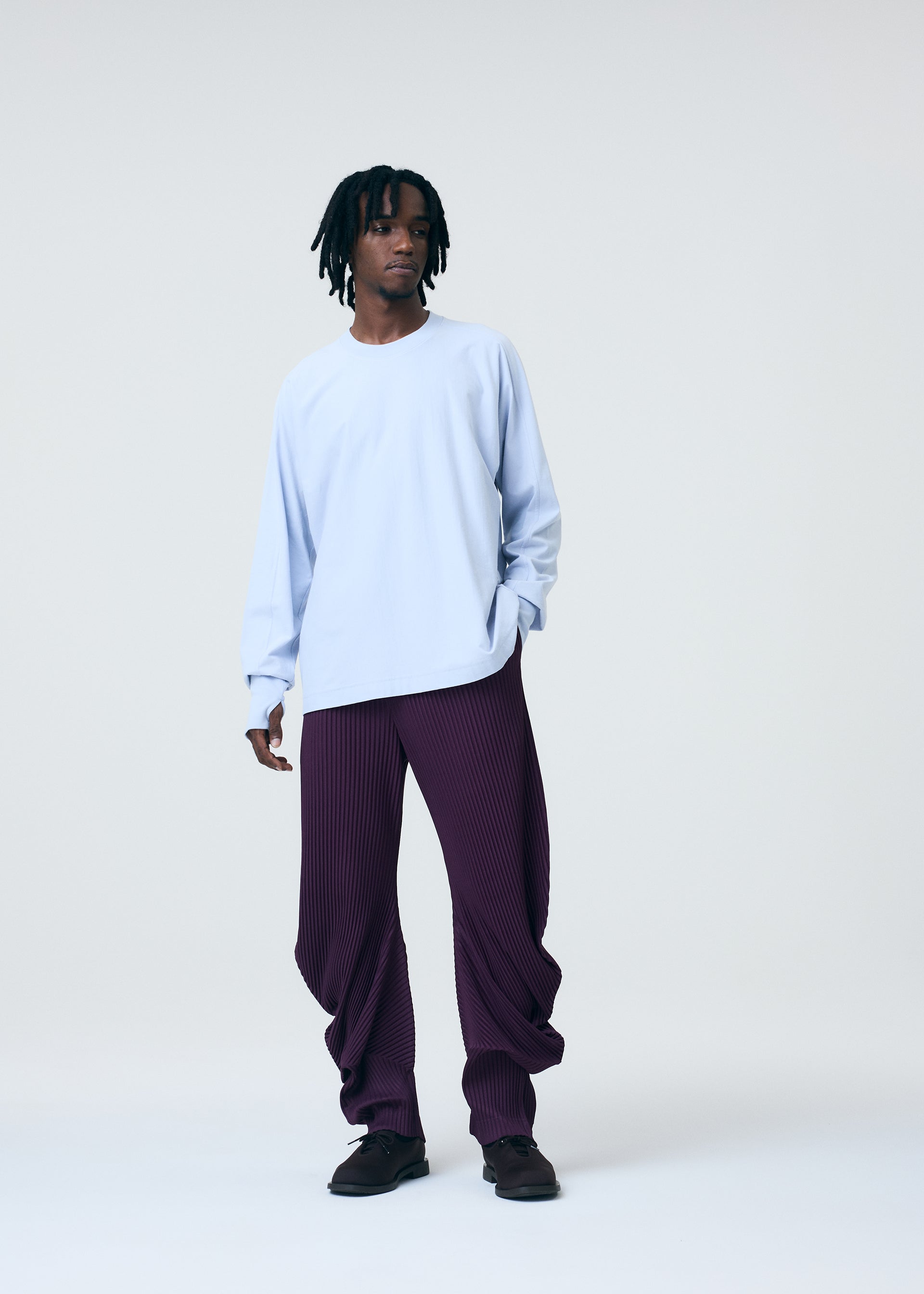 Zoomed view A man with short dreadlocks stands against a white background, wearing the HOMME PLISSÉ ISSEY MIYAKE RELEASE-T 1 cotton top with thumbholes, textured baggy purple pants, and dark shoes.