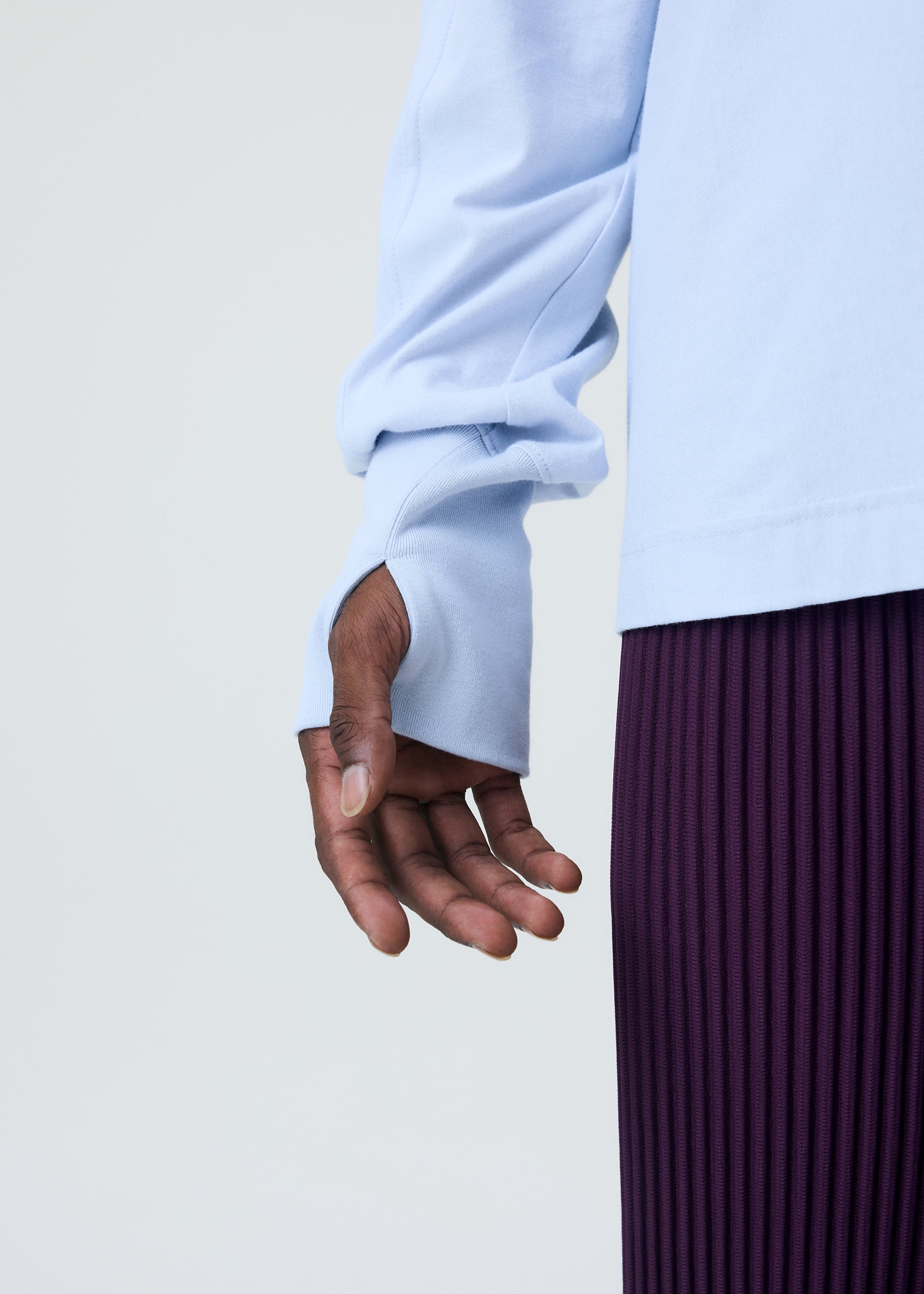 Zoomed view A person wearing the light blue RELEASE-T 1 cotton round-neck long-sleeved top with thumbholes by HOMME PLISSÉ ISSEY MIYAKE and a pleated purple skirt, hand relaxed at their side against a plain light background.