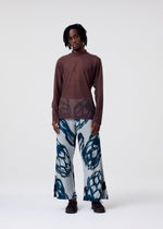 A man with short, twisted hair stands against a plain white background, wearing HOMME PLISSÉ ISSEY MIYAKE’s CRYSTALLINE JERSEY—a brown sheer top with semi-sheer panels and wide-leg pants featuring bold blue and black abstract patterns.