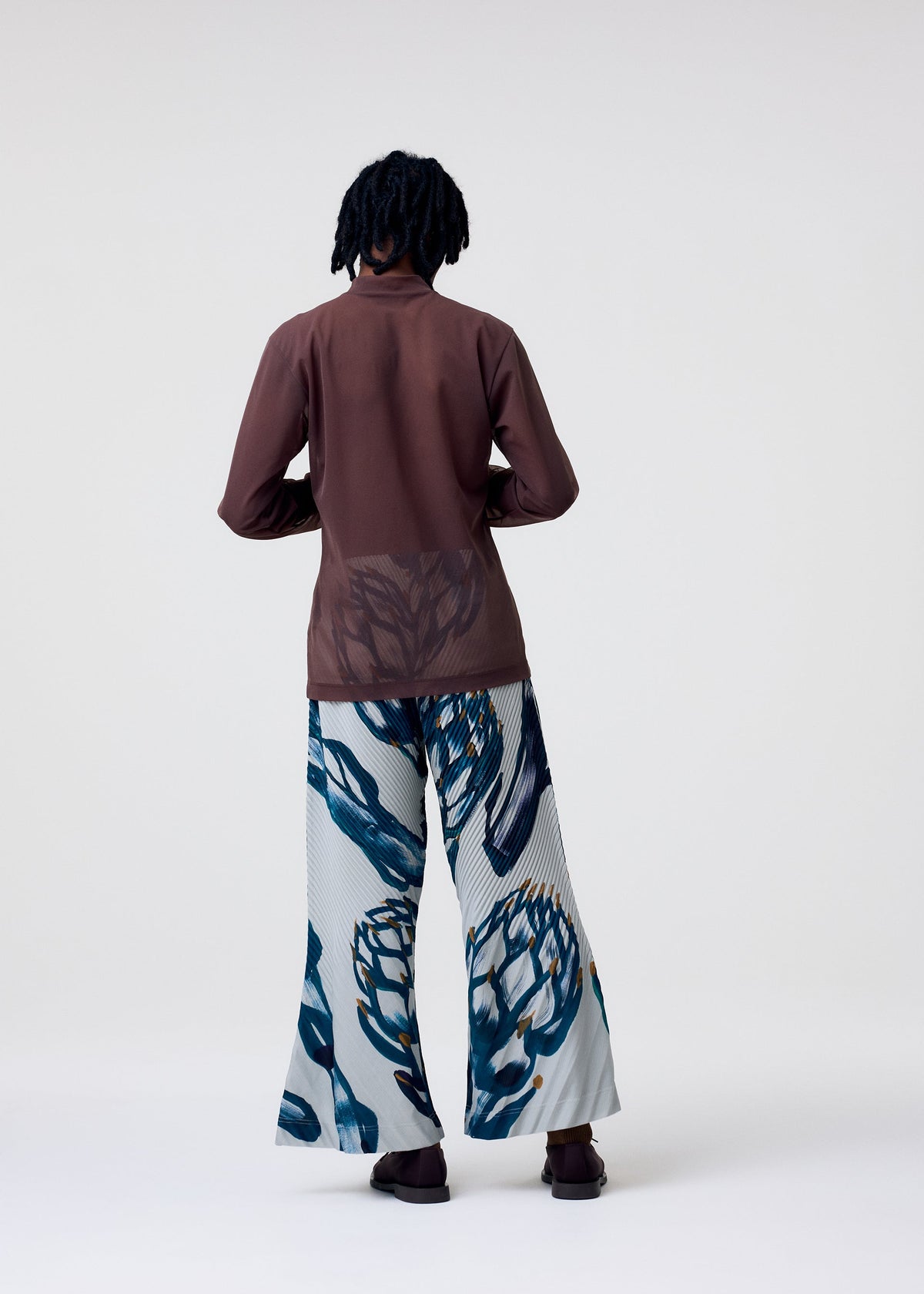 A person stands with their back to the camera, wearing the HOMME PLISSÉ ISSEY MIYAKE CRYSTALLINE JERSEY: a sheer maroon mock neck top and wide-leg pants with an abstract blue and white floral pattern, set against a plain light background. view 5 of 5