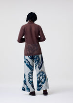 A person stands with their back to the camera, wearing the HOMME PLISSÉ ISSEY MIYAKE CRYSTALLINE JERSEY: a sheer maroon mock neck top and wide-leg pants with an abstract blue and white floral pattern, set against a plain light background.