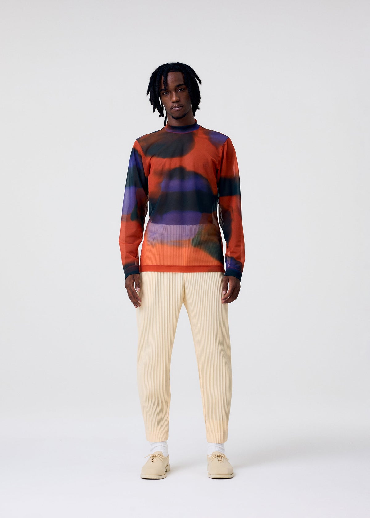Wearing the DAWN SHEER mock-neck top by HOMME PLISSÉ ISSEY MIYAKE, a person with medium-length dreadlocks pairs it with cream ribbed pants, white socks, and beige shoes against a plain light background. view 4 of 8
