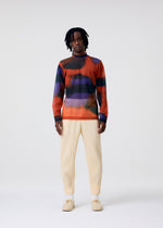 Wearing the DAWN SHEER mock-neck top by HOMME PLISSÉ ISSEY MIYAKE, a person with medium-length dreadlocks pairs it with cream ribbed pants, white socks, and beige shoes against a plain light background.