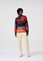 A person with medium-length dreadlocks wears the DAWN SHEER top by HOMME PLISSÉ ISSEY MIYAKE, cream ribbed pants, and beige shoes, standing against a plain light gray background.
