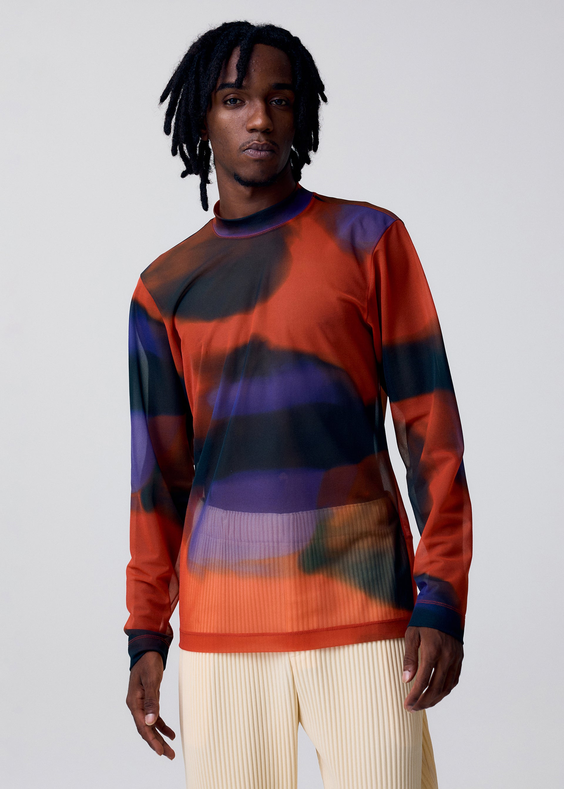 Zoomed view A person with medium-length dreadlocks wears the HOMME PLISSÉ ISSEY MIYAKE DAWN SHEER mock-neck top, featuring a bold orange, black, and purple abstract pattern, paired with cream pleated pants against a plain light background.