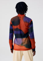 A person with medium-length dreadlocks stands facing away, wearing the DAWN SHEER mock-neck top by HOMME PLISSÉ ISSEY MIYAKE—its bold red, purple, and black abstract pattern paired with light pleated pants against a plain background.