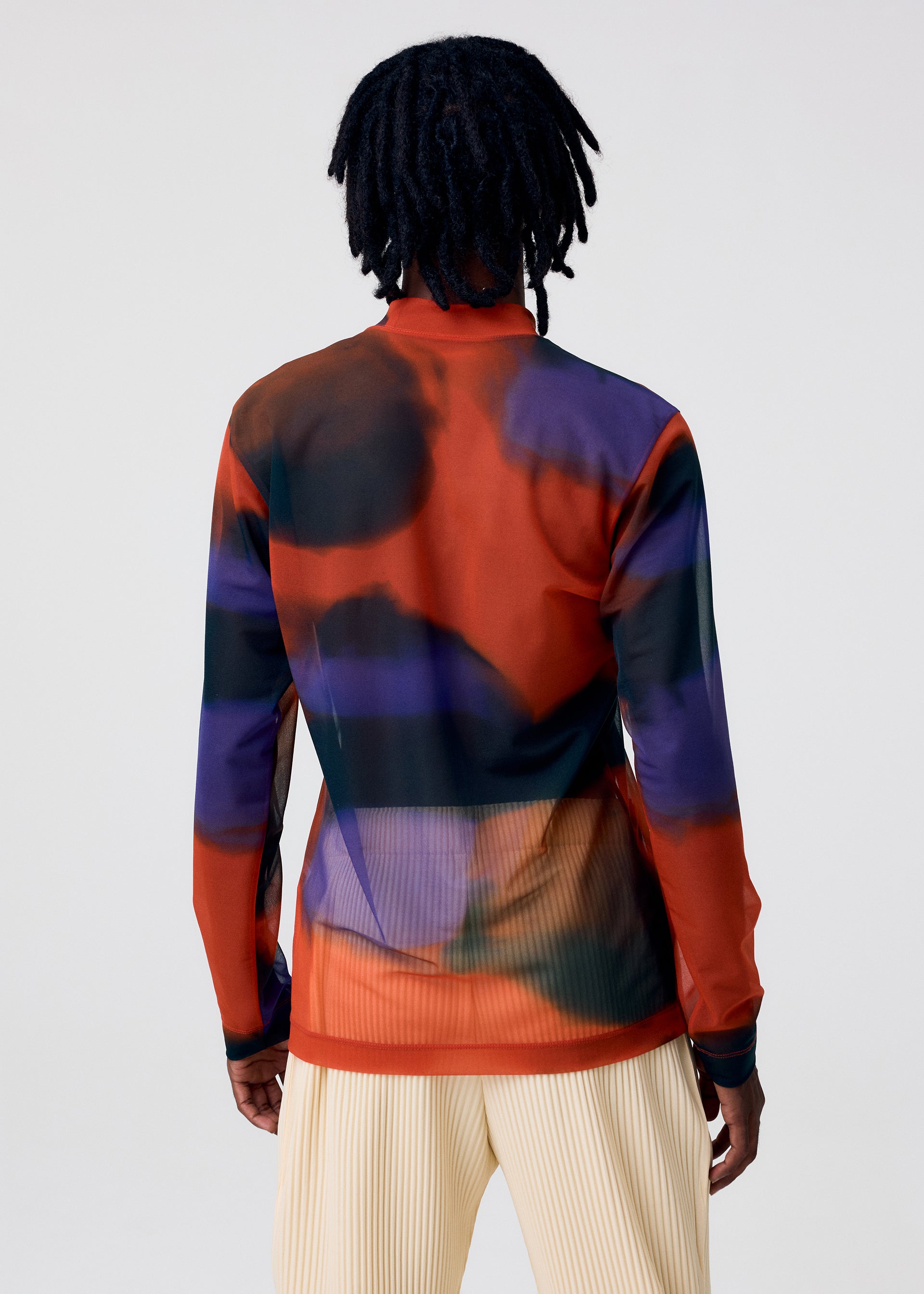Zoomed view A person with medium-length dreadlocks stands facing away, wearing the DAWN SHEER mock-neck top by HOMME PLISSÉ ISSEY MIYAKE—its bold red, purple, and black abstract pattern paired with light pleated pants against a plain background.