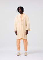 A person with shoulder-length dark hair and a neutral expression wears the HOMME PLISSÉ ISSEY MIYAKE MONTHLY COLORS : APRIL set—an airy cream kimono jacket over a light orange shirt and pants, styled with white socks and beige shoes against a plain background.