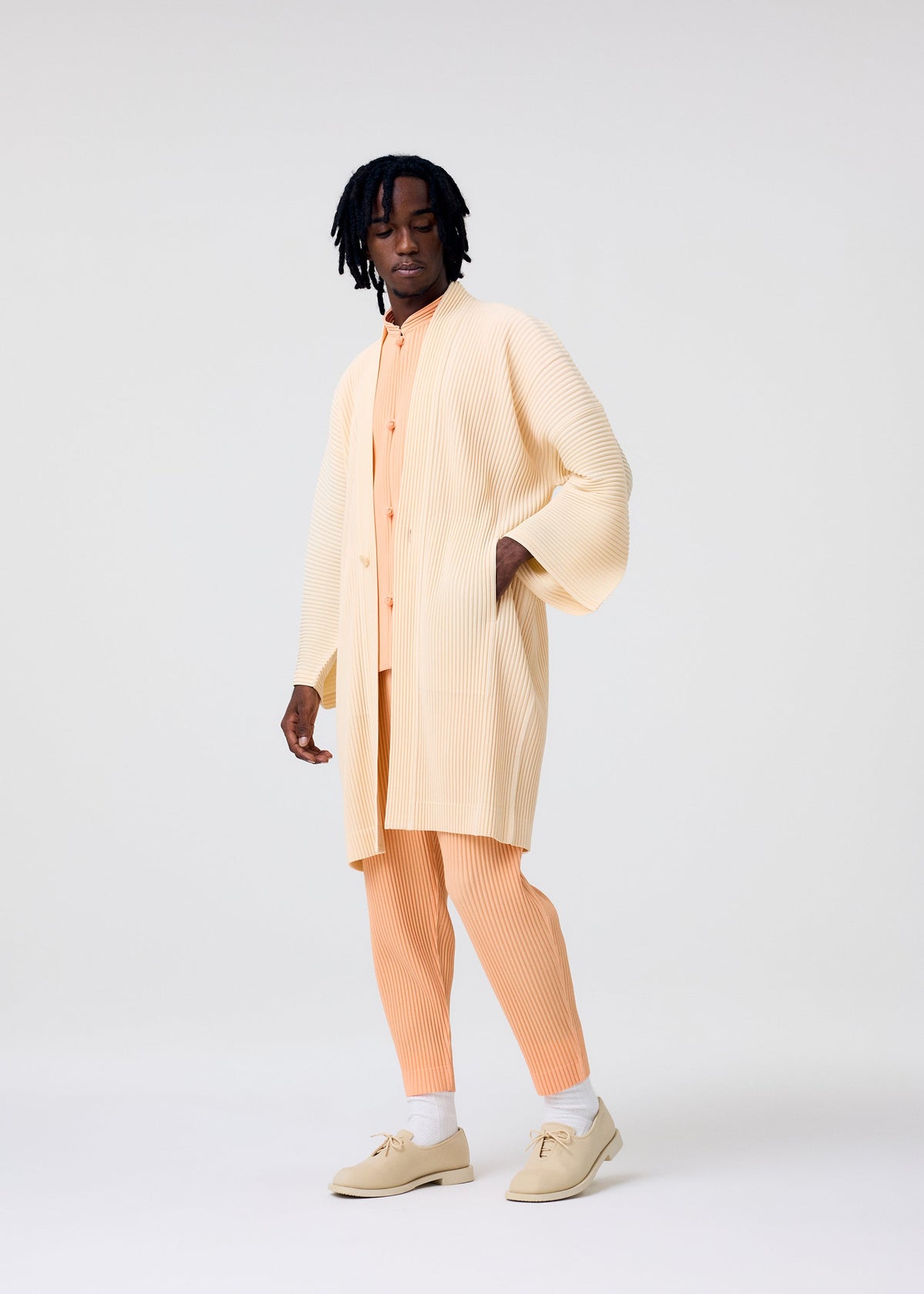 A person with medium-dark skin tone and short dreadlocks wears pieces from the HOMME PLISSÉ ISSEY MIYAKE MONTHLY COLORS : APRIL collection—cream kimono-style pleated overcoat, peach shirt and pants, and beige shoes—against a light background. view 5 of 6
