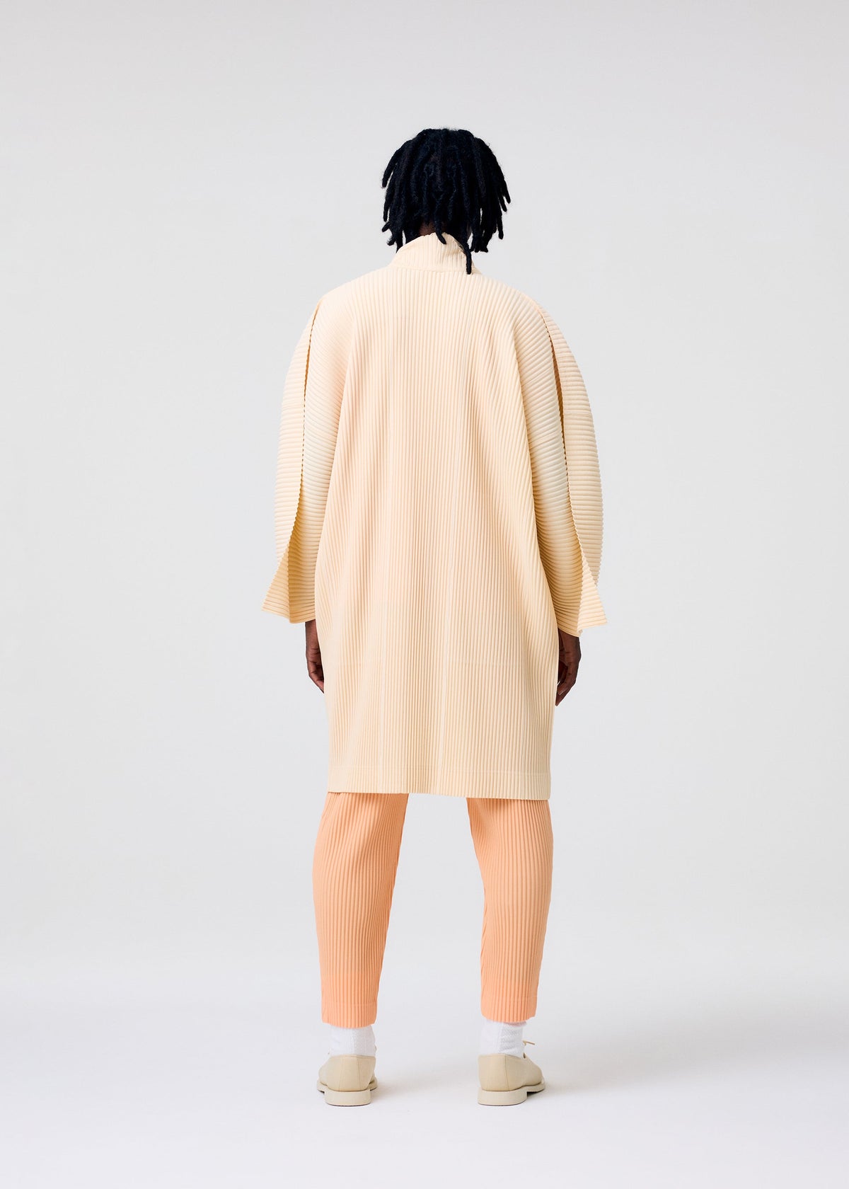 A person with medium-length dreadlocks stands facing away in HOMME PLISSÉ ISSEY MIYAKE's MONTHLY COLORS : APRIL—a cream kimono-style pleated coat, peach pleated pants, and cream slip-on shoes—against a plain light background. view 6 of 6
