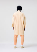 A person with medium-length dreadlocks stands facing away in HOMME PLISSÉ ISSEY MIYAKE's MONTHLY COLORS : APRIL—a cream kimono-style pleated coat, peach pleated pants, and cream slip-on shoes—against a plain light background.