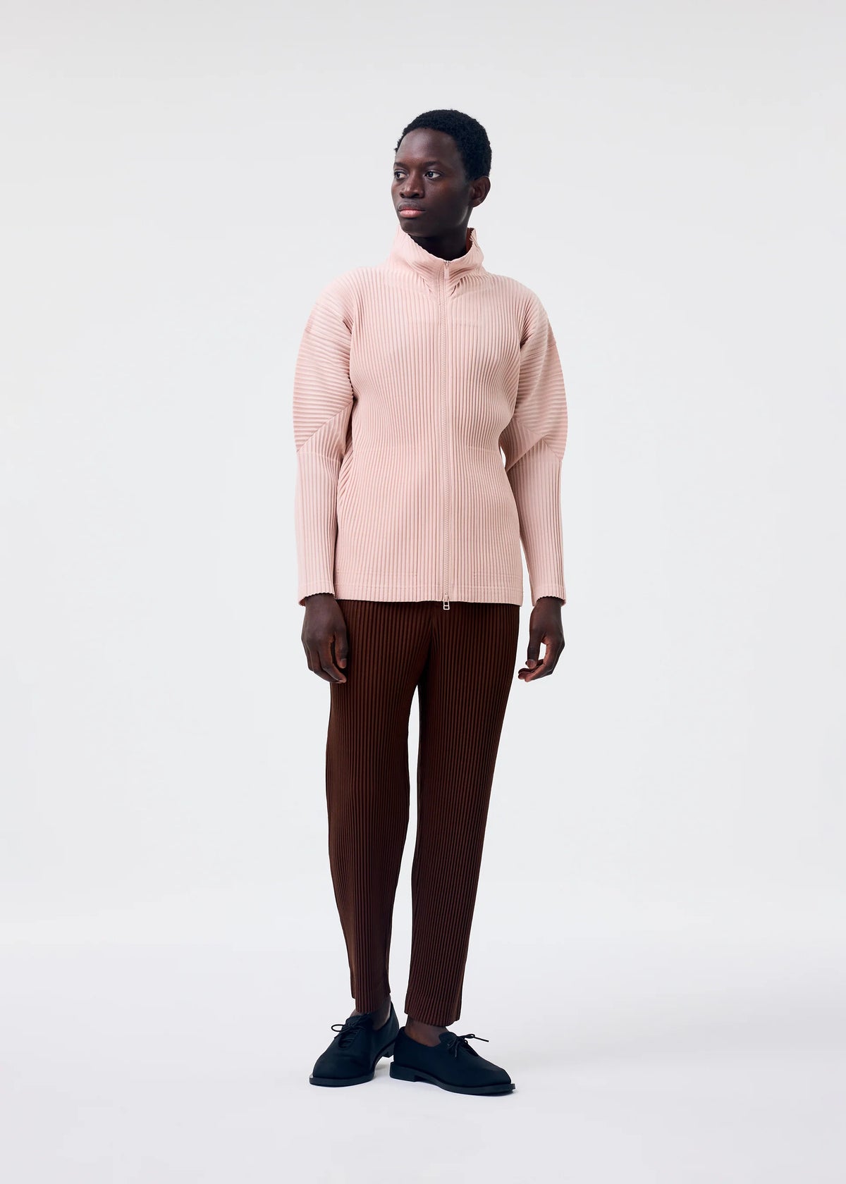 A person stands against a light background, wearing a pale pink COLOR PLEATS 2 zip-up sweatshirt by HOMME PLISSÉ ISSEY MIYAKE, dark brown ribbed pants, and black shoes, looking slightly to the side. view 6 of 6