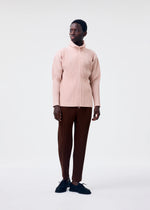 A person stands against a light background, wearing a pale pink COLOR PLEATS 2 zip-up sweatshirt by HOMME PLISSÉ ISSEY MIYAKE, dark brown ribbed pants, and black shoes, looking slightly to the side.