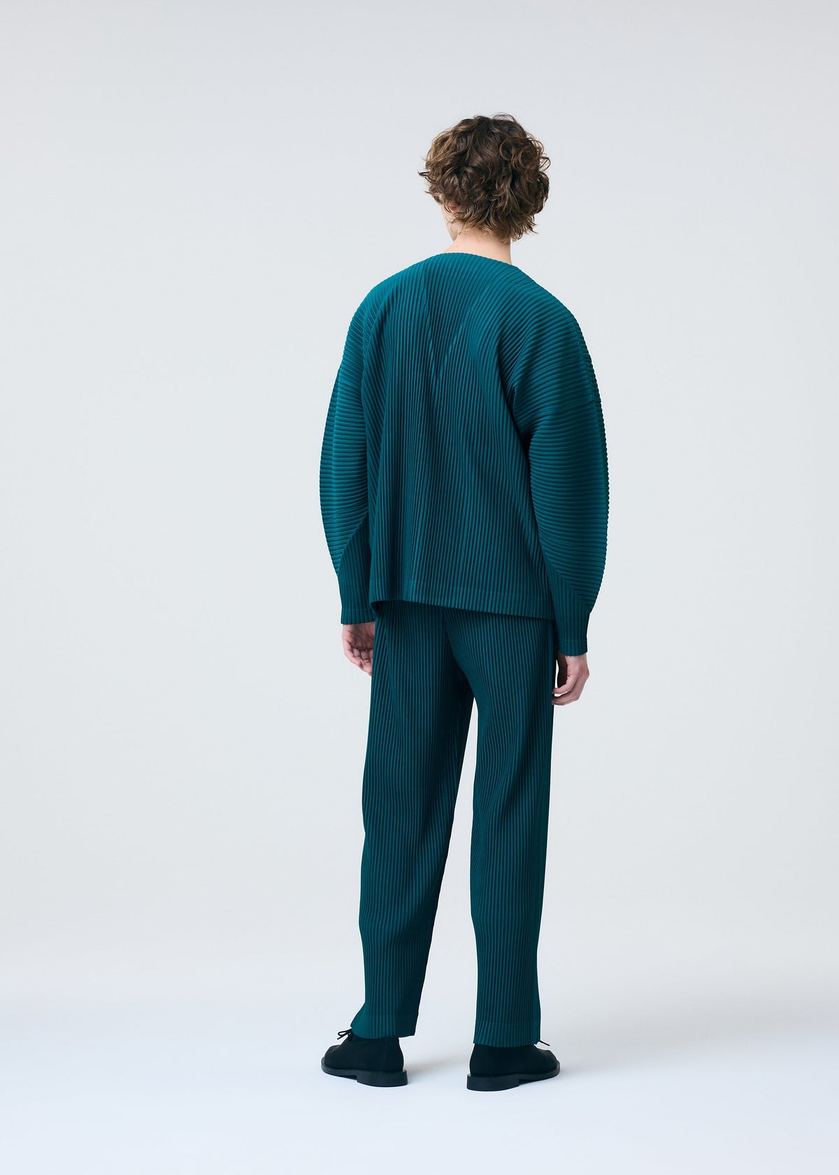 A person with short, curly hair faces away in a loose-fitting teal pinstriped COLOR PLEATS 1 cardigan and pants by HOMME PLISSÉ ISSEY MIYAKE, paired with black shoes, set against a plain light background. view 7 of 7