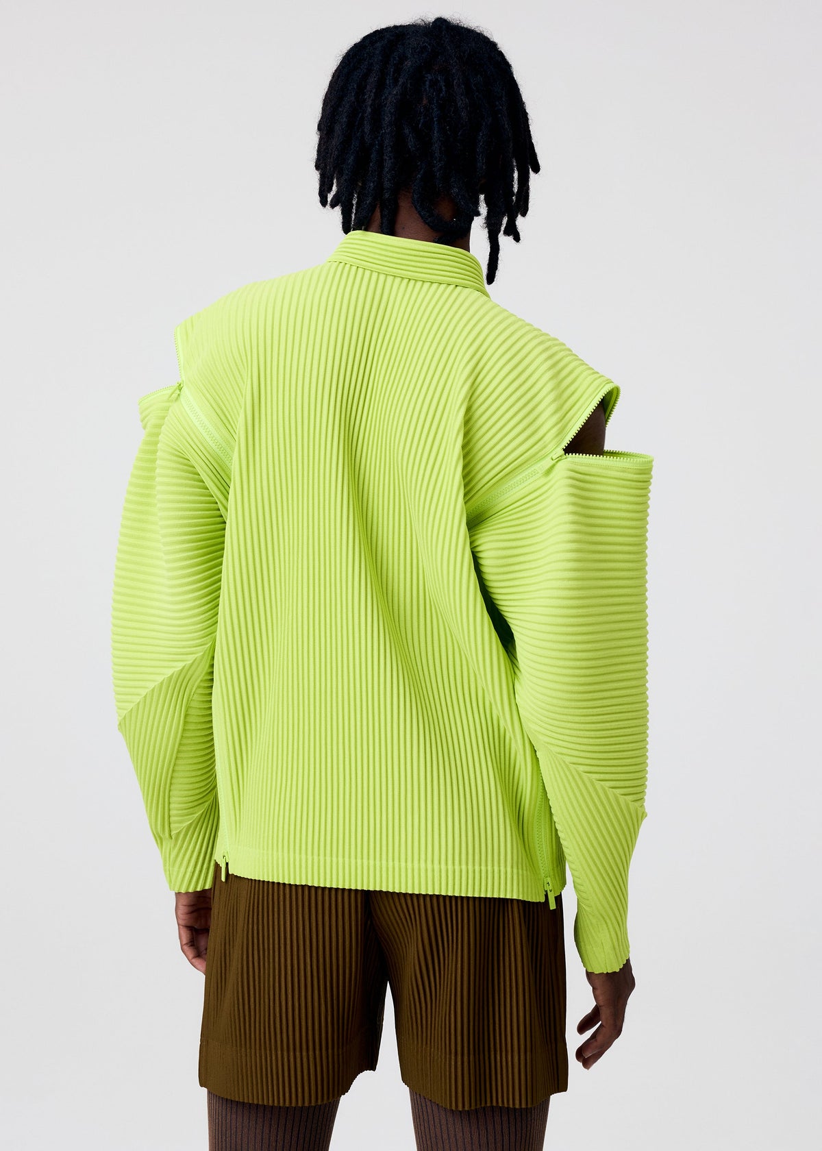 A person with short dreadlocks is seen from the back wearing the HOMME PLISSÉ ISSEY MIYAKE MULTI-FASTENED yellow-green textured jacket with cutout shoulders and brown pleated shorts, set against a plain light background. view 6 of 7