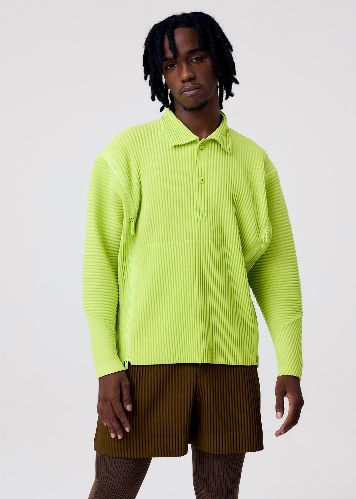 A person with short dreadlocks wears the HOMME PLISSÉ ISSEY MIYAKE MULTI-FASTENED lime green, long-sleeved polo shirt and brown ribbed shorts against a plain light background, showcasing versatile styling for any occasion. view 7 of 7